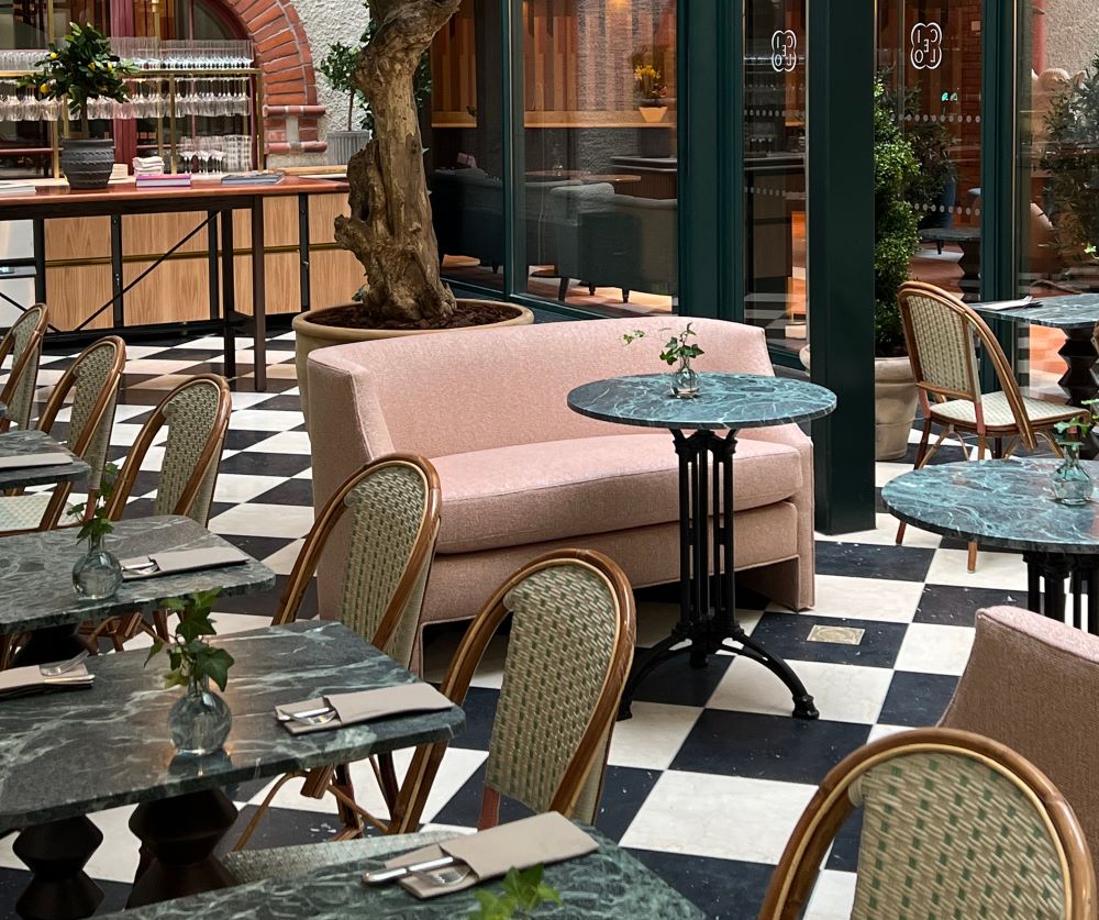Lounge- och restaurangmiljö med bistrokänsla. Svart/vit rutigt marmorgolv, rosa soffa, bistrobord i gjutjärn med gröna marmorskivor samt handflätade bistrostolar.